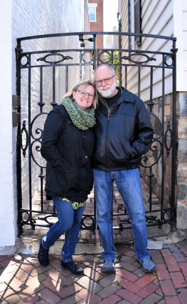 Man with white beard and leather jacket wearing blue jeans standing close to a woman with mid-length blonde hair. She has a black jacket and green heavy scarf on. They are standing on a brick sidewalk in front of a wrought iron gate. Terry and Nancy